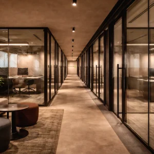 A modern office hallway with glass-walled meeting rooms on both sides, warm lighting, and a neutral color palette; lounge chairs and tables are visible in the foreground—expert office interior design and fit out by interioritalia.