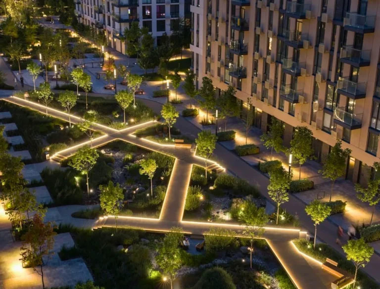 A modern apartment complex at night with illuminated walkways forming geometric patterns among landscaped greenery and small trees, creating a vibrant and inviting outdoor space.