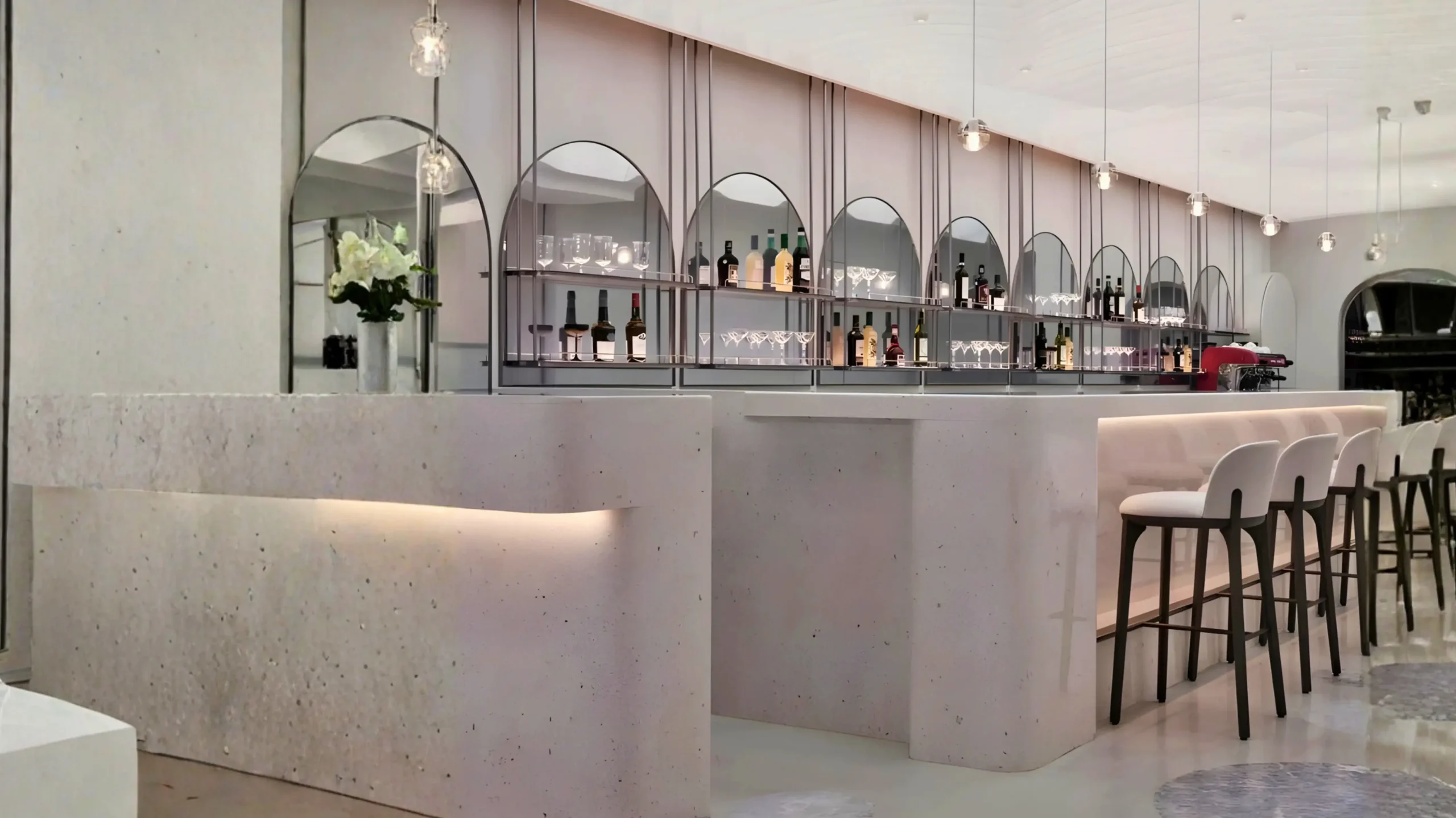 Modern bar interior with a curved counter, high stools, and a back wall displaying bottles and glassware under arched mirrors, with pendant lights hanging from the ceiling.
