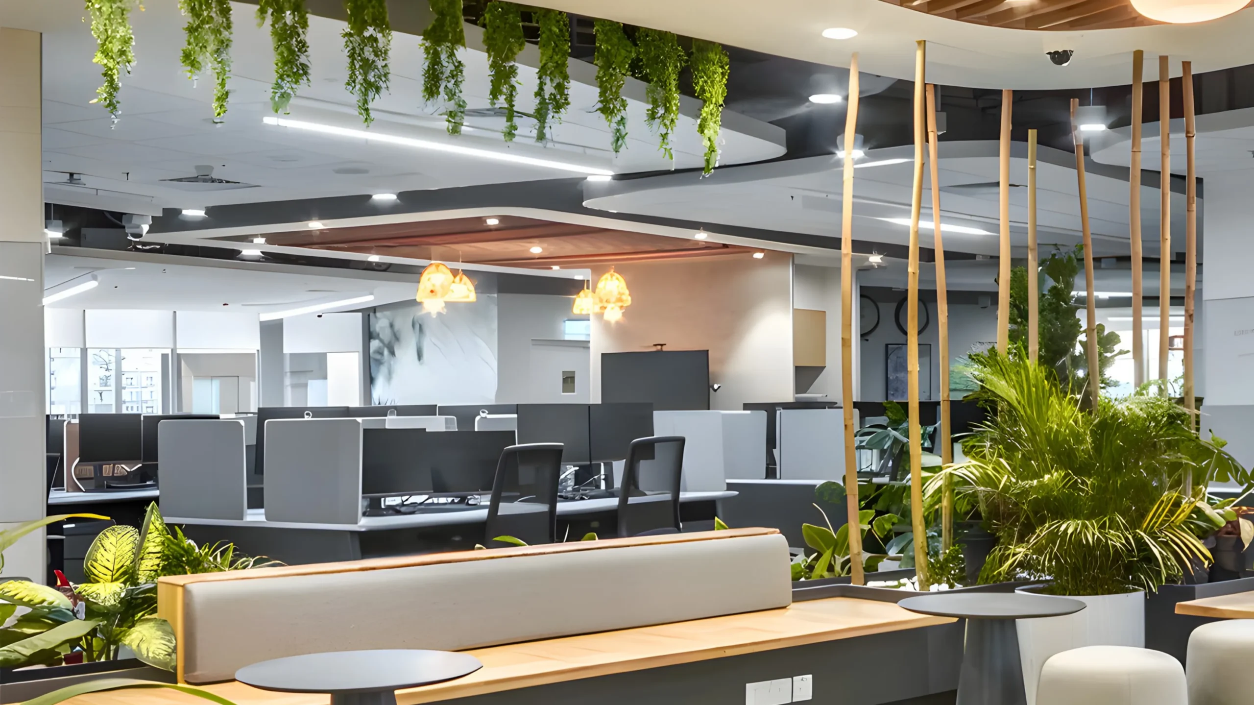 Modern open-plan office with gray desks, black chairs, and hanging plants. The space features greenery, wooden accents, and natural light, creating a fresh, inviting work environment with a collaborative seating area in the foreground.